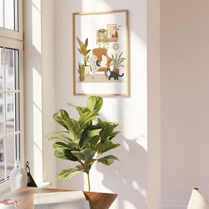 May include: A framed art print featuring a cozy interior scene with two cats. The artwork depicts a brown armchair, plants, and decorative items. The cats are black and white. The frame is a light wood color. The print is displayed on a white wall.