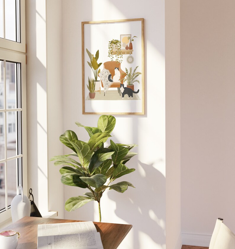 May include: A framed art print featuring a cozy interior scene with two cats. The artwork depicts a brown armchair, plants, and decorative items. The cats are black and white. The frame is a light wood color. The print is displayed on a white wall.