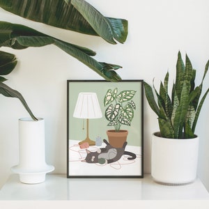 May include: A framed art print featuring a cat playing with yarn, a lamp, and a potted plant. The artwork has a light green background and a black frame. The cat is gray and black. The print is displayed on a white surface with plants in the background.