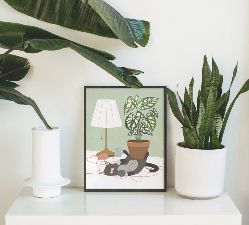 May include: A framed art print featuring a cat playing with yarn, a lamp, and a potted plant. The artwork has a light green background and a black frame. The cat is gray and black. The print is displayed on a white surface with plants in the background.