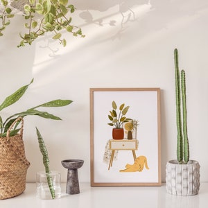 May include: A framed art print featuring a stylized illustration of a potted plant on a small table, a yellow cat stretching, and other decorative plants. The print is in a light wood frame. A tall cactus is in a concrete pot.