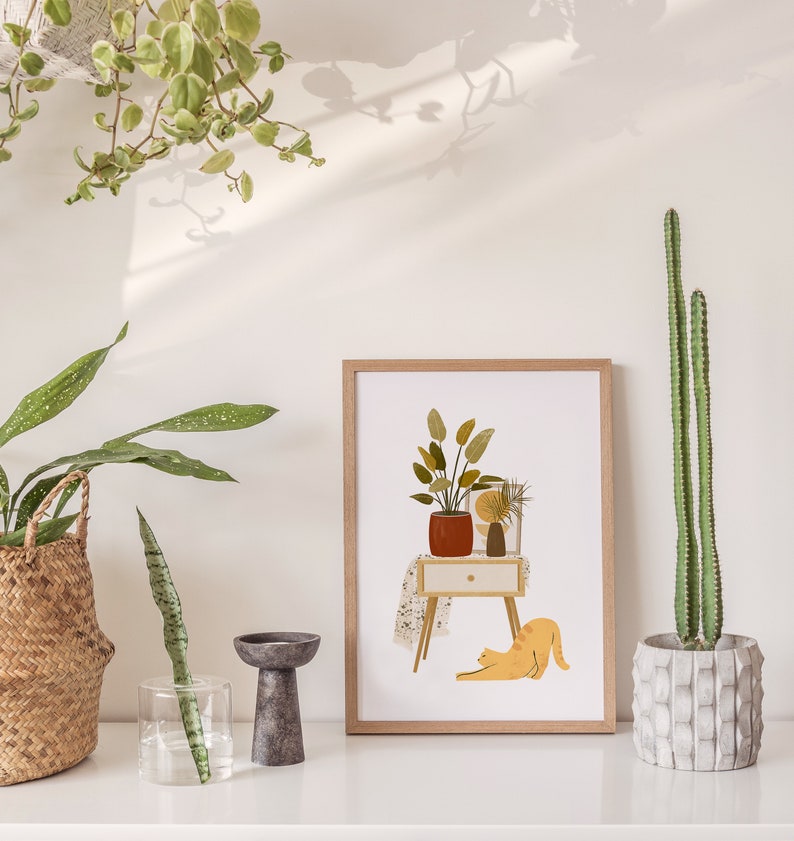 May include: A framed art print featuring a stylized illustration of a potted plant on a small table, a yellow cat stretching, and other decorative plants. The print is in a light wood frame. A tall cactus is in a concrete pot.