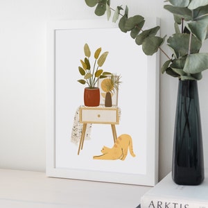 May include: A framed art print featuring a stylized illustration of a potted plant, a small table with a drawer, and a stretching orange cat. A dark vase with greenery is on the right. The artwork has a minimalist, modern aesthetic.