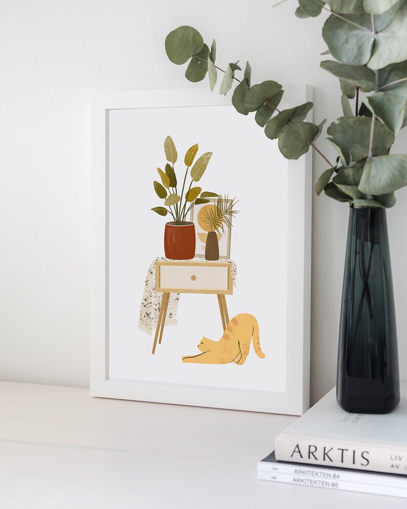 May include: A framed art print featuring a stylized illustration of a potted plant, a small table with a drawer, and a stretching orange cat. A dark vase with greenery is on the right. The artwork has a minimalist, modern aesthetic.