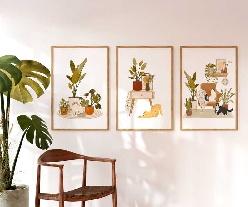 May include: Three framed art prints featuring illustrations of cats and plants. One print shows a cat sitting among potted plants. Another depicts a cat stretching near a nightstand with a plant. The third print shows cats on a chair surrounded by plants.