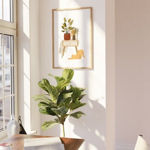 May include: A framed art print featuring a minimalist illustration of a nightstand with plants and a cat. The artwork has a warm color palette with shades of orange, brown, and green. The frame is a light wood color, and the print is displayed on a white wall.