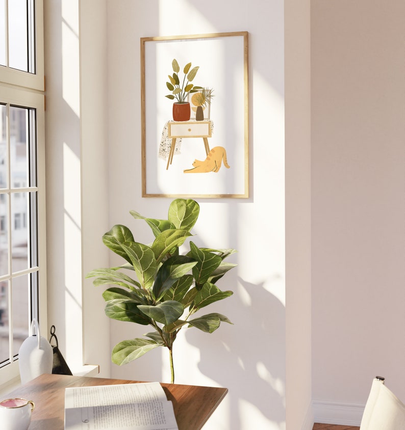 May include: A framed art print featuring a minimalist illustration of a nightstand with plants and a cat. The artwork has a warm color palette with shades of orange, brown, and green. The frame is a light wood color, and the print is displayed on a white wall.