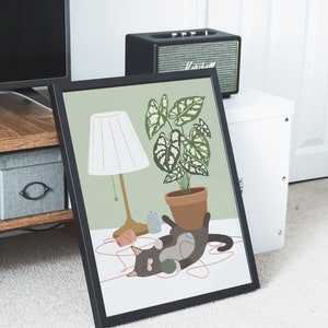 May include: Framed art print featuring a playful cat with a ball of yarn, a potted plant, and a lamp. The illustration has a soft color palette of green, brown, and cream. The cat is dark gray with white markings.