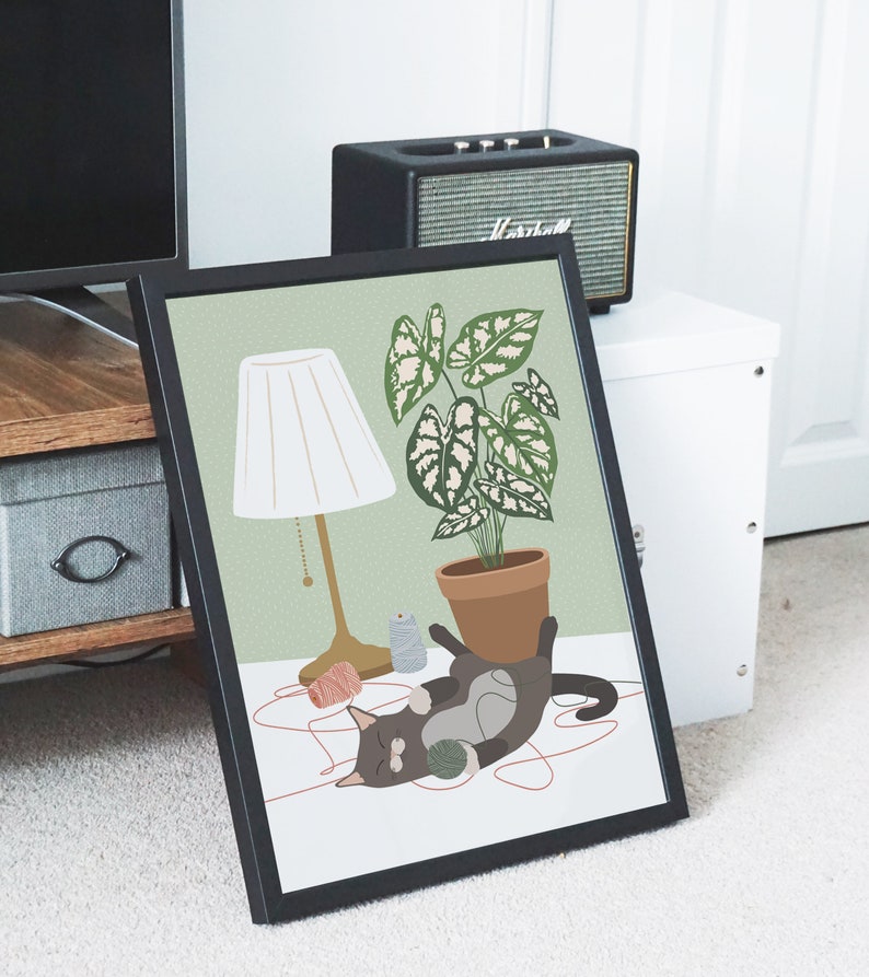 May include: Framed art print featuring a playful cat with a ball of yarn, a potted plant, and a lamp. The illustration has a soft color palette of green, brown, and cream. The cat is dark gray with white markings.