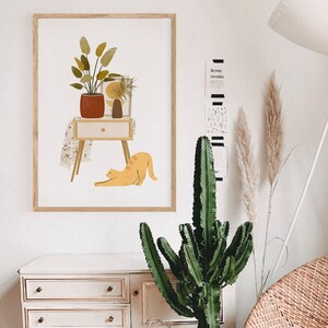 May include: A framed art print featuring a mid-century modern illustration. The artwork depicts a potted plant, a small table with a drawer, a vase, and a stretching orange cat. The color palette includes shades of orange, brown, and green.