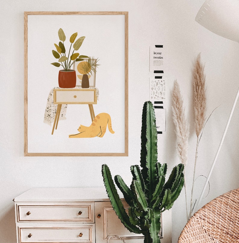 May include: A framed art print featuring a mid-century modern illustration. The artwork depicts a potted plant, a small table with a drawer, a vase, and a stretching orange cat. The color palette includes shades of orange, brown, and green.