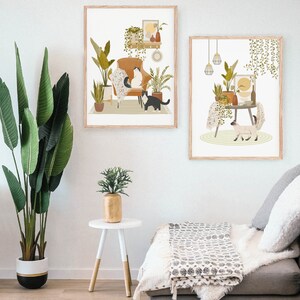 May include: Two framed art prints featuring illustrations of cats in cozy indoor settings. One print shows cats on a chair surrounded by plants, the other shows a cat near a table with plants. The prints have a neutral color palette with wooden frames.