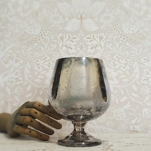 May include: A silver-toned metal goblet with a wide, rounded bowl and a short, sturdy stem. The surface of the goblet features a weathered, tarnished finish with visible dark patches and a reflective, metallic sheen throughout the exterior.