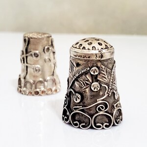 May include: Two silver thimbles with intricate floral designs. The thimbles are both round and have a smooth, shiny finish.