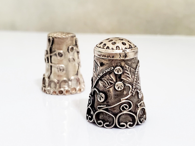 May include: Two silver thimbles with intricate floral designs. The thimbles are both round and have a smooth, shiny finish.