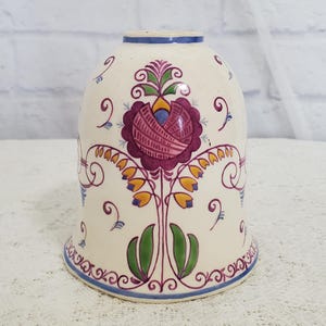 May include: Cream-colored ceramic lampshade with a floral design. The design features a large, stylized purple flower at the center, surrounded by green leaves, yellow and orange accents, and blue and purple swirls. The rim is trimmed with a blue border.