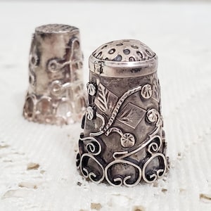 May include: Two silver thimbles with intricate floral designs. The thimbles are made of metal and have a shiny finish.