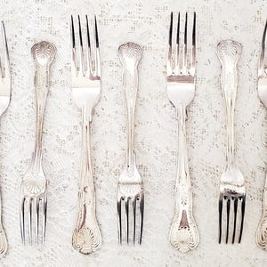 May include: An assortment of silver forks with ornate, detailed handles. The forks are arranged on a white, textured surface, showcasing their elegant design. The tines are sharp and the handles feature floral and scrollwork patterns.