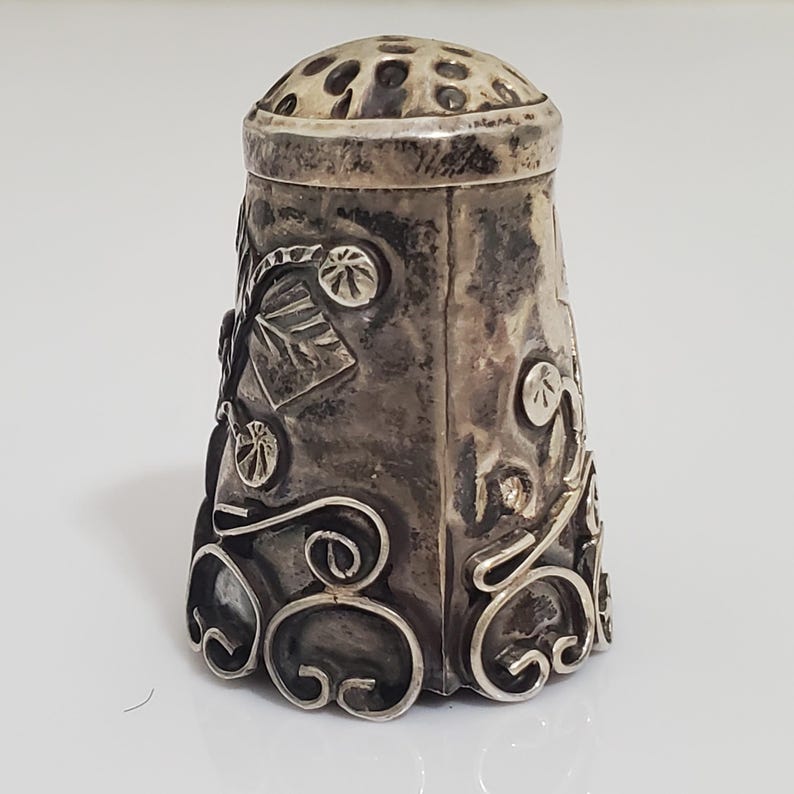 May include: A silver thimble with an ornate design featuring floral motifs and swirls. The thimble has a textured top with small holes.