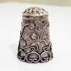 May include: A silver thimble with an intricate floral design. The thimble has a textured surface and a small hole on the top.
