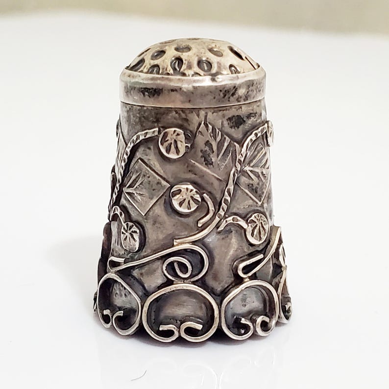 May include: A silver thimble with an intricate floral design. The thimble has a textured surface and a small hole on the top.