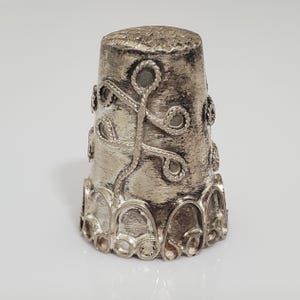 May include: A silver thimble with an intricate design of a stylized plant with leaves and stems. The thimble has a rounded top and a scalloped base.