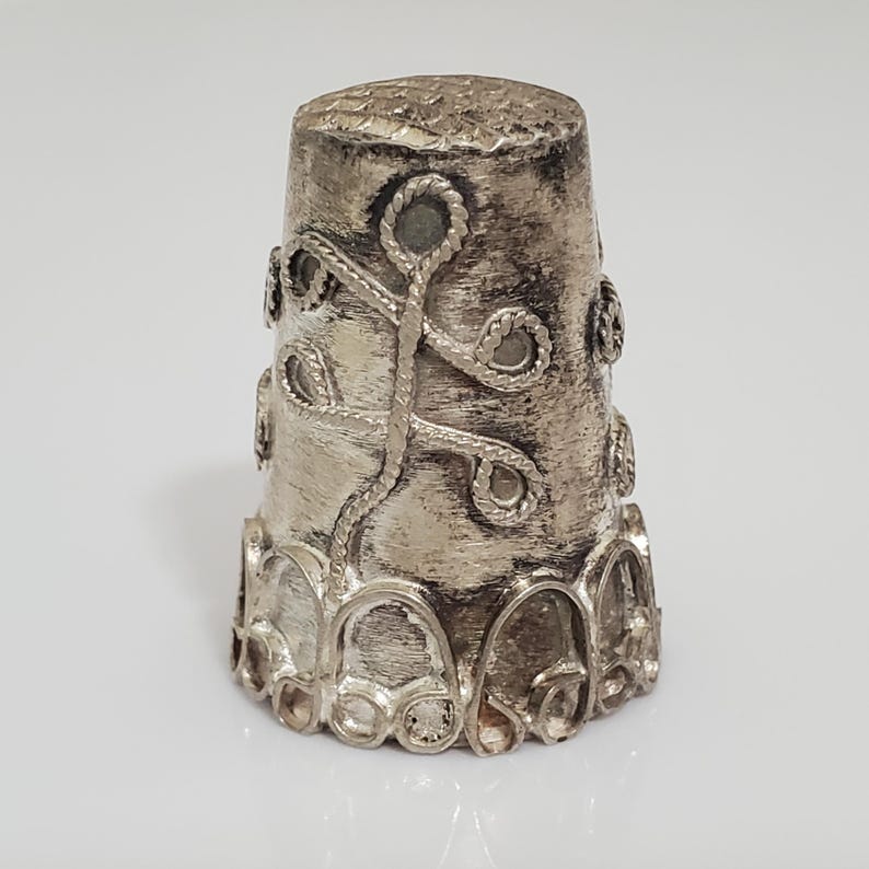 May include: A silver thimble with an intricate design of a stylized plant with leaves and stems. The thimble has a rounded top and a scalloped base.