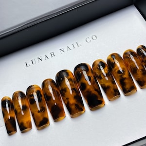 May include: A set of ten tortoise shell press-on nails in a long, coffin shape. The nails are arranged in a row in a white box with the text "LUNAR NAIL CO" printed on the box.