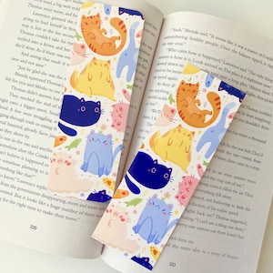 May include: Two colorful bookmarks featuring a repeating pattern of cute cartoon cats in various colors and poses. The cats are surrounded by stars and fish. The bookmarks are perfect for book lovers who want to add a touch of whimsy to their reading experience.
