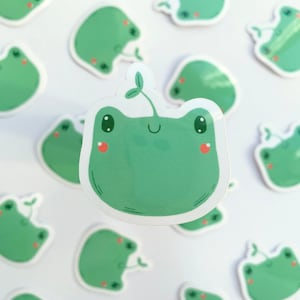May include: A selection of green frog stickers, each with a sprout detail. The stickers depict a smiling frog face with rosy cheeks and large eyes. The stickers are set against a white backdrop.