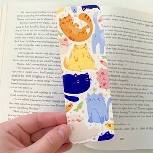 May include: A white bookmark with a repeating pattern of colorful cats. The cats are in various poses and have different expressions. The bookmark is decorated with small flowers and stars.