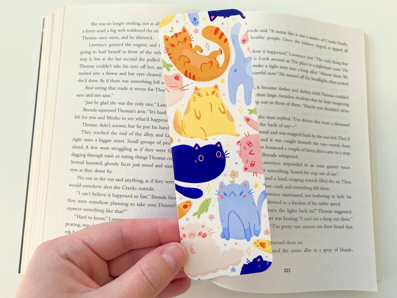 May include: A white bookmark with a repeating pattern of colorful cats. The cats are in various poses and have different expressions. The bookmark is decorated with small flowers and stars.