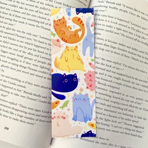 May include: A colorful bookmark featuring a variety of cartoon cats in different poses and colors. The cats are on a white background with a few small flowers and fish. The bookmark is perfect for book lovers who enjoy whimsical designs.