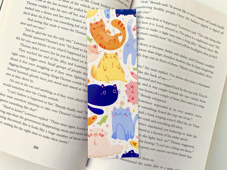 May include: A colorful bookmark featuring a variety of cartoon cats in different poses and colors. The cats are on a white background with a few small flowers and fish. The bookmark is perfect for book lovers who enjoy whimsical designs.