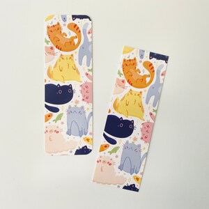 May include: Two white bookmarks with a repeating pattern of colorful cats. The cats are in various poses, including sitting, lying down, and standing. The background is white with small stars and fish.