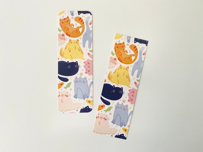 May include: Two white bookmarks with a repeating pattern of colorful cats. The cats are in various poses, including sitting, lying down, and standing. The background is white with small stars and fish.