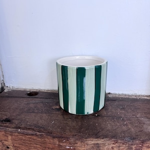 May include: A cylindrical ceramic planter featuring vertical stripes in alternating shades of dark forest green and light sage green. The interior of the vessel is a smooth, solid off-white colour, and the exterior has a glossy, reflective finish.
