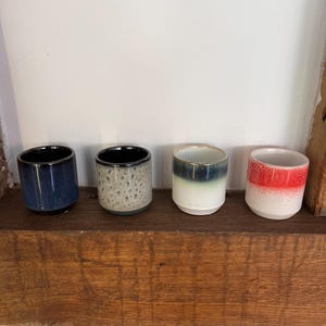 May include: Four ceramic cups with a rounded shape and a glossy finish. The cups are arranged on a wooden shelf. Each cup has a different colour scheme: navy blue, speckled grey, white with green and blue, and white with red.