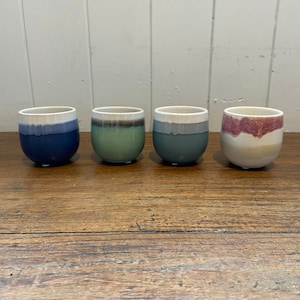 May include: Four ceramic cups with a rounded shape and a two-tone colour scheme. The cups are in shades of blue, green, and red, with a white rim. The cups are arranged on a wooden surface.