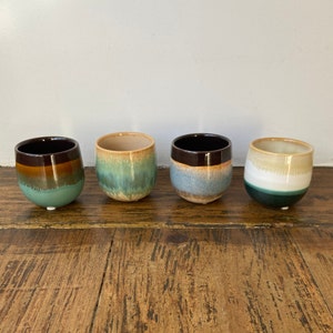 May include: Four ceramic stemless cups with a rounded shape. Each cup features a unique colour scheme, including shades of brown, green, blue, and white. The cups are arranged on a wooden surface, showcasing their varied glazed finishes.
