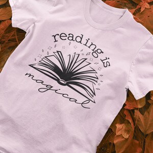 May include: A pale pink t-shirt featuring the phrase "reading is magical" in black script, accompanied by an open book illustration. The shirt is set against a backdrop of autumnal leaves.