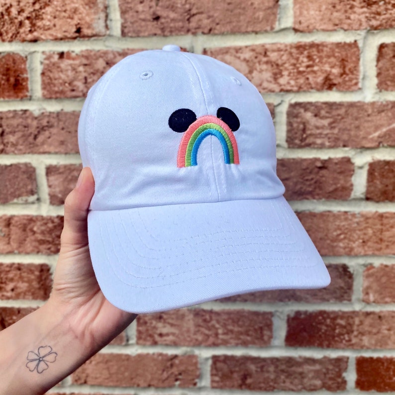May include: White baseball cap with a rainbow and black mouse ears embroidered on the front.