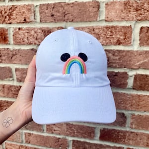 May include: White baseball cap with a rainbow and Mickey Mouse ears embroidered on the front.