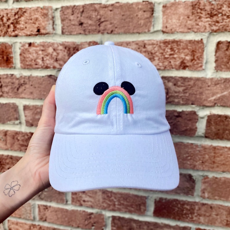 May include: White baseball cap with a rainbow and Mickey Mouse ears embroidered on the front.