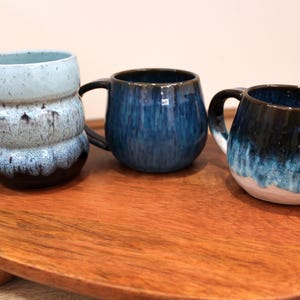 May include: Three ceramic mugs with distinctive blue and white glaze patterns. The mugs have rounded bodies and curved handles. The mugs are displayed on a wooden serving tray. The mugs are suitable for coffee, tea, or other beverages.