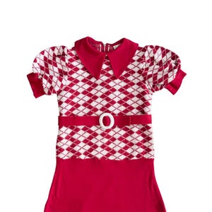 May include: A red and white argyle pattern dress with a white belt and buckle. The dress has a red collar and short sleeves. The bottom of the dress is solid red. A vintage style dress.