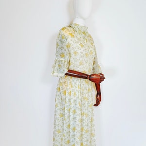 May include: A white maxi dress with a floral print in shades of yellow and green. The dress has a high neck, short sleeves with ruffles, and a gathered skirt.