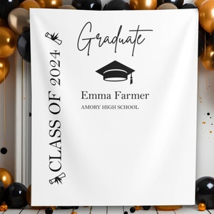 Puede incluir: Telón de fondo de graduación blanco con el texto "Graduate" y "Class of 2024". Incluye un gráfico de gorro de graduación y el nombre "Emma Farmer" con "Amory High School" debajo. Globos negros y dorados en el fondo.