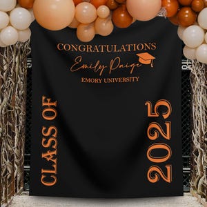 May include: Black graduation banner with orange text that reads "CONGRATULATIONS Emily Paige EMORY UNIVERSITY" and "CLASS OF 2025". The banner is decorated with balloons in shades of orange and white.