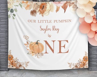 Può includere: Striscione in tessuto bianco con illustrazioni floreali e di zucche. Il testo recita "OUR LITTLE PUMPKIN Saylor Ray is ONE". Lo striscione ha un tema autunnale con colori arancione, marrone e crema. Palloncini nei toni del pesca e crema sono a destra.